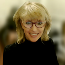 Head and shoulders image of a blond woman wearing a black turtleneck and reading glasses.